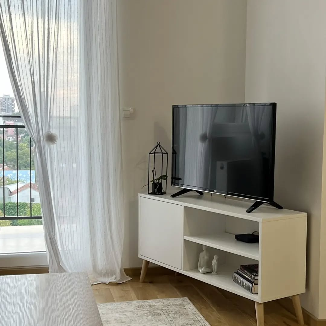 Modern living room corner with a black flat-screen TV on a white wooden media console, sheer white curtains, and a small potted plant in a black geometric holder.