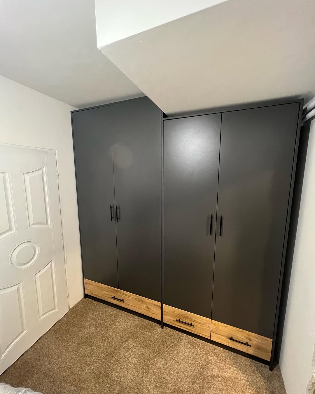 Two dark gray wardrobes with black handles and wooden drawer fronts placed side by side in a carpeted room corner near a white door.