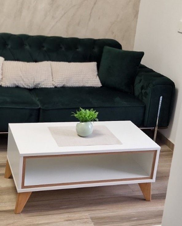 Modern living room with a dark green velvet tufted sofa adorned with white and dark green cushions, and a white rectangular coffee table with wooden legs and a small green potted plant on top.