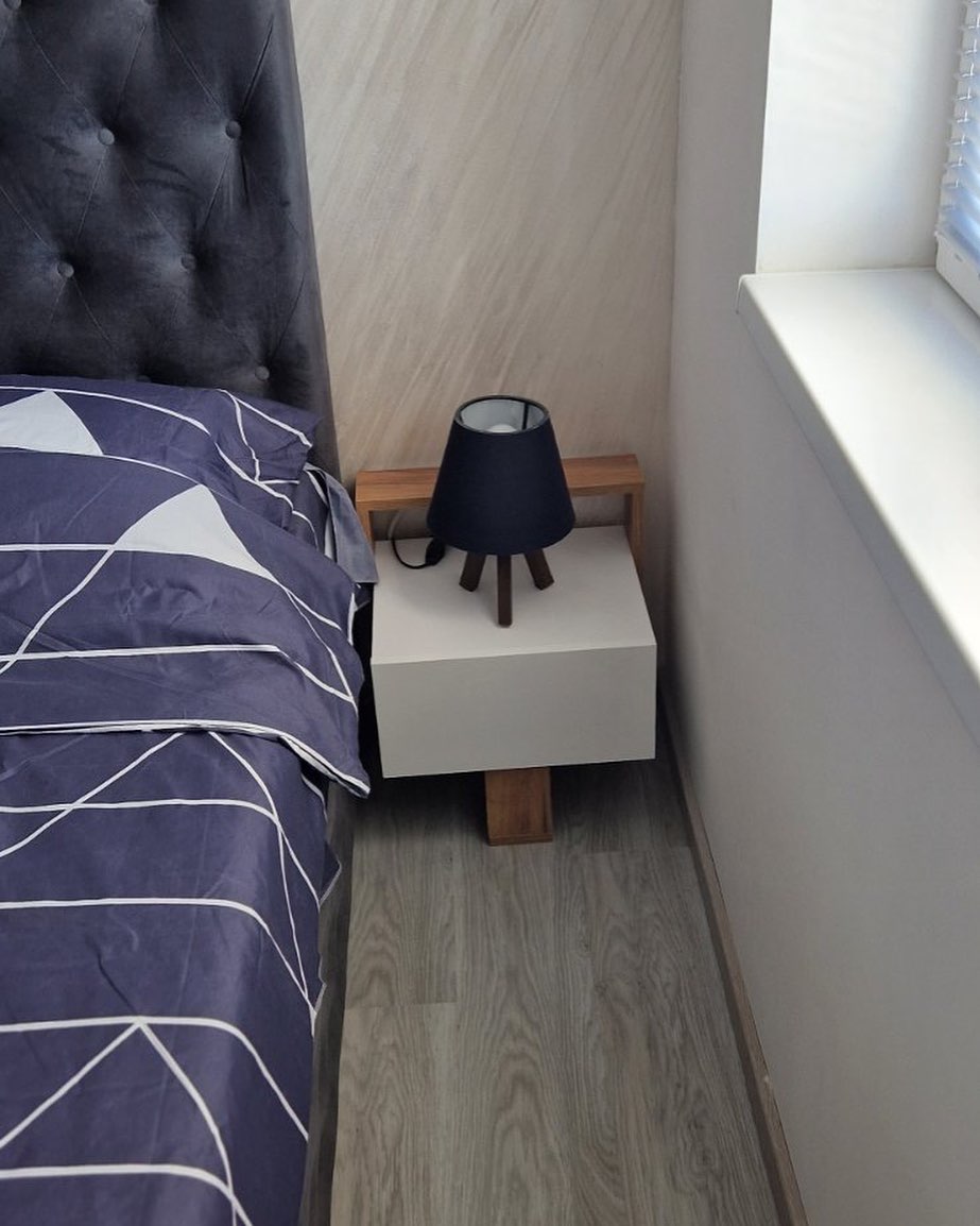 Bed with dark tufted headboard next to a white nightstand holding a small dark blue lamp near a window with blinds.