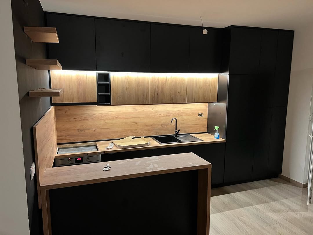 Modern kitchen with black and wood cabinetry, black sink with faucet, under-cabinet lighting, oven, and a wooden breakfast bar with a key and some dust on top.