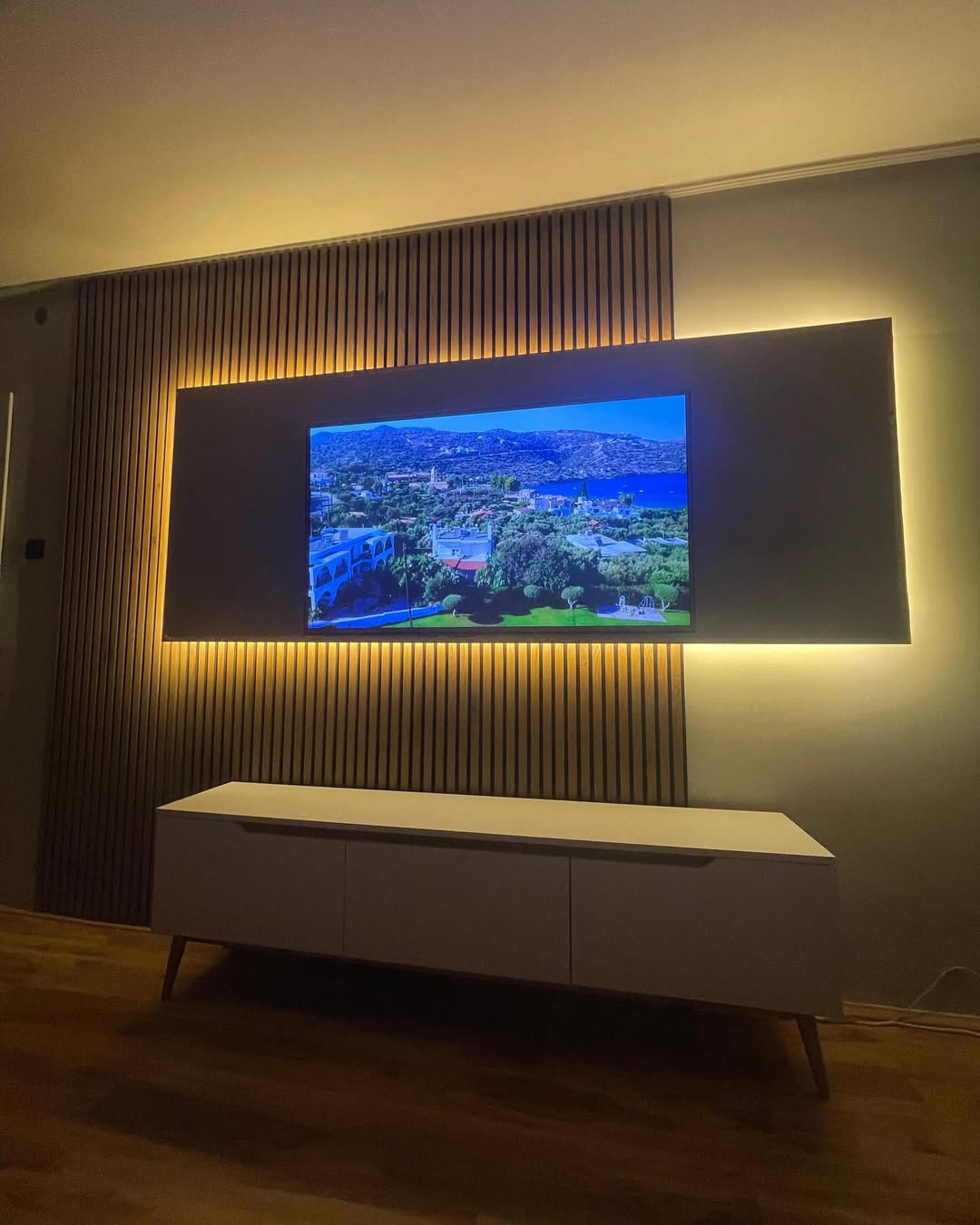 Wall-mounted flat-screen TV with backlighting displaying a scenic coastal landscape above a modern white cabinet.