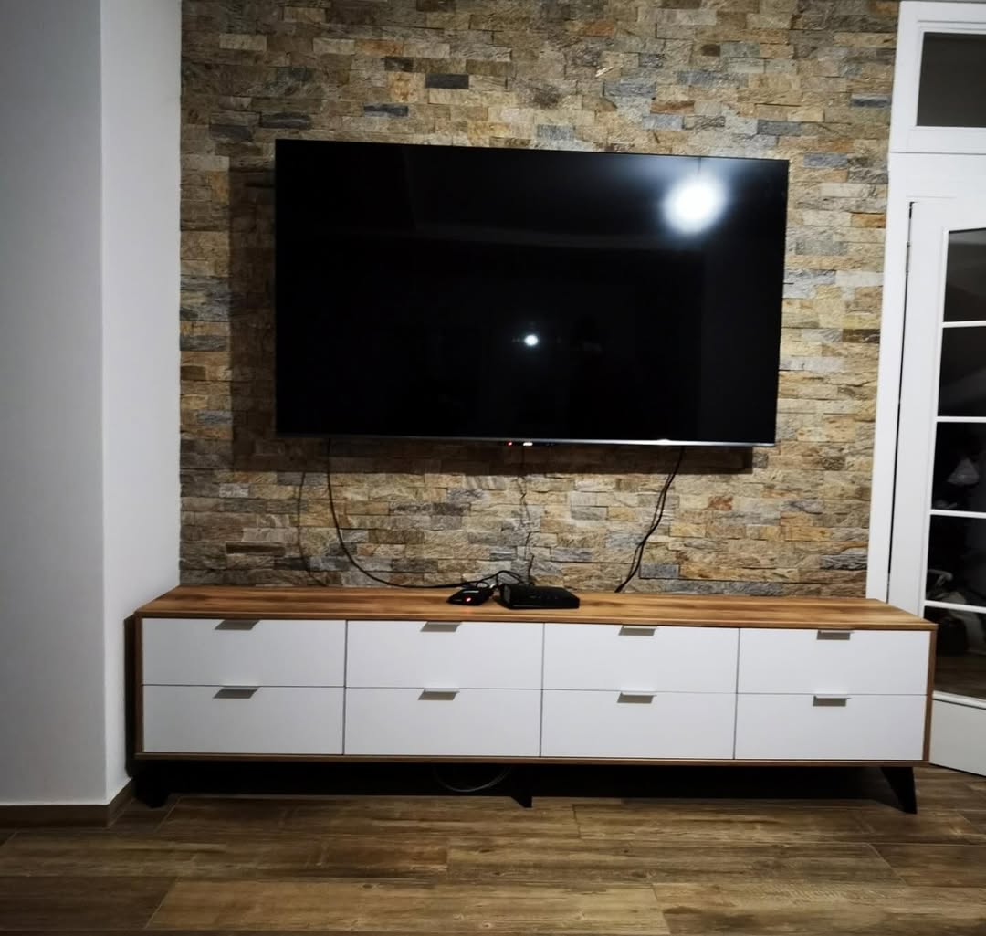 Flat-screen TV mounted on a stone brick wall above a modern wooden and white cabinet with electronic devices on top.