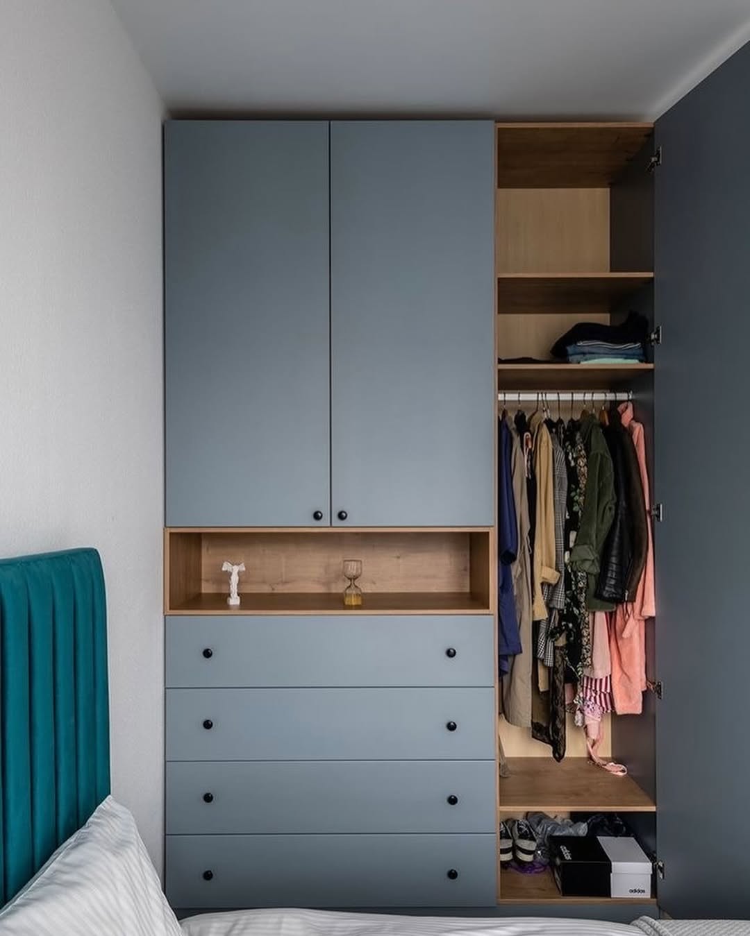 Blue wardrobe with one open door showing hanging clothes, shelves with folded clothes, shoes, and boxes beside a bed with teal headboard.