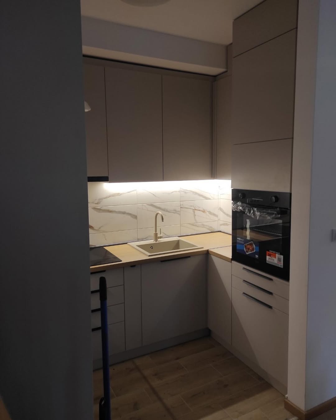 Compact modern kitchen with beige cabinets, a built-in oven, sink, and marble-patterned backsplash under under-cabinet lighting.